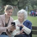 Delighted female relatives sitting together on wooden bench in park and browsing mobile phone while learning using