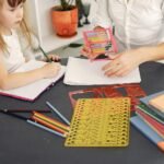 From above of little girl sitting at table with school items and textbooks while writing in notebook with pencil together with unrecognizable young home teacher in white shirt with abacus in hand during early home education