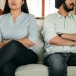 Crop faceless young ethnic couple in casual wear sitting on couch with arms crossed keeping silence after scandal at home