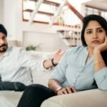 Young bearded Sikh man in casual wear and turban arguing with sad wife sitting on sofa