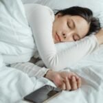 Calm Asian female wearing white pajama sleeping in comfortable bed with white sheets near modern mobile phone in morning